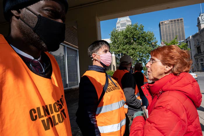 An unmasked woman stands in front of a masked person wearing an orange jacket identifying them as a clinic escort volunteer