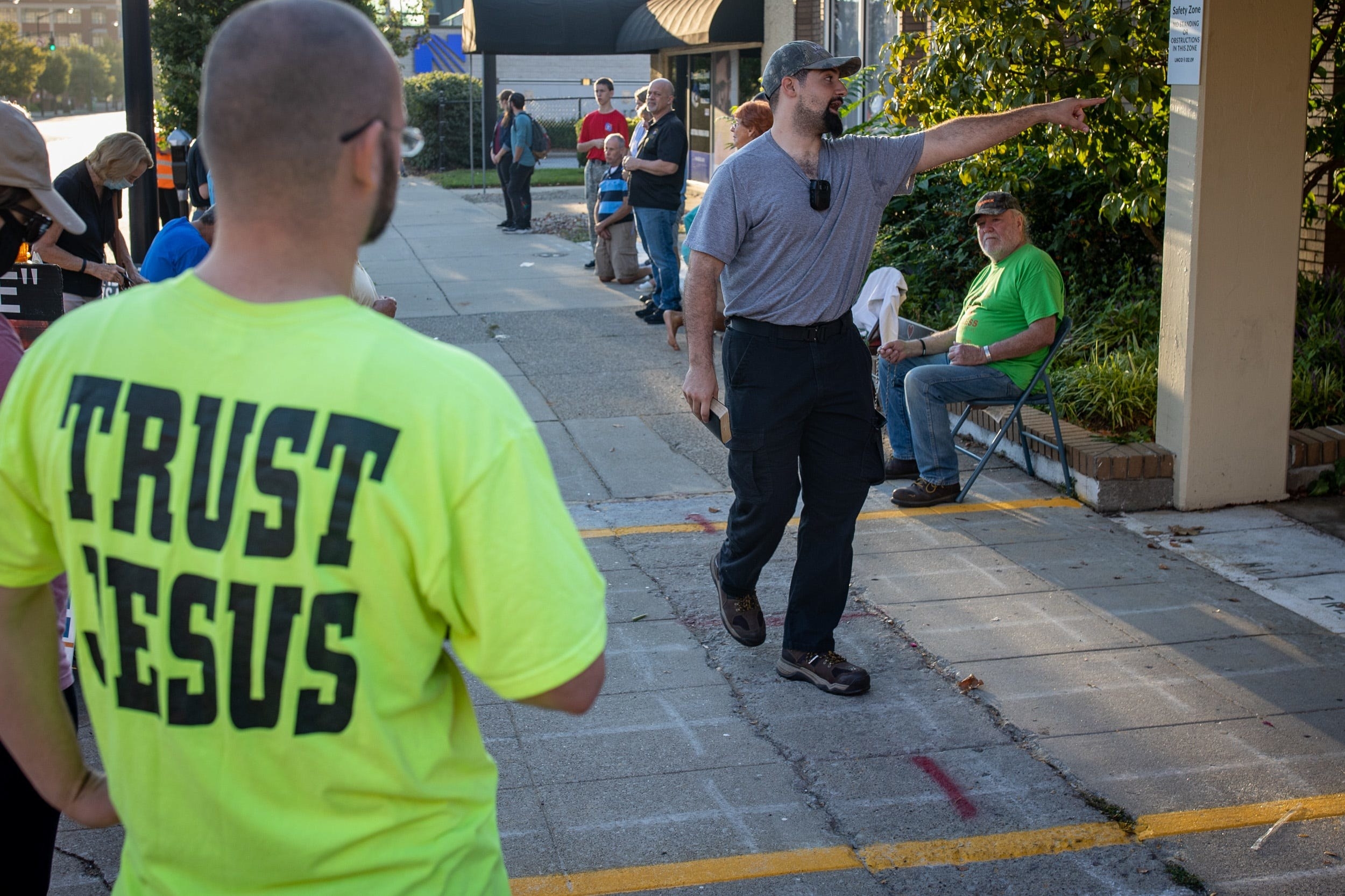 A person with &quot;Trust Jesus&quot; on the back of their T-shirt watches the protester