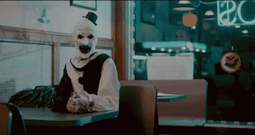 A creepy clown putting his head on his hands, smiling, in a diner