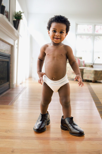 A baby walks around the living room in their dad&#x27;s shoes.