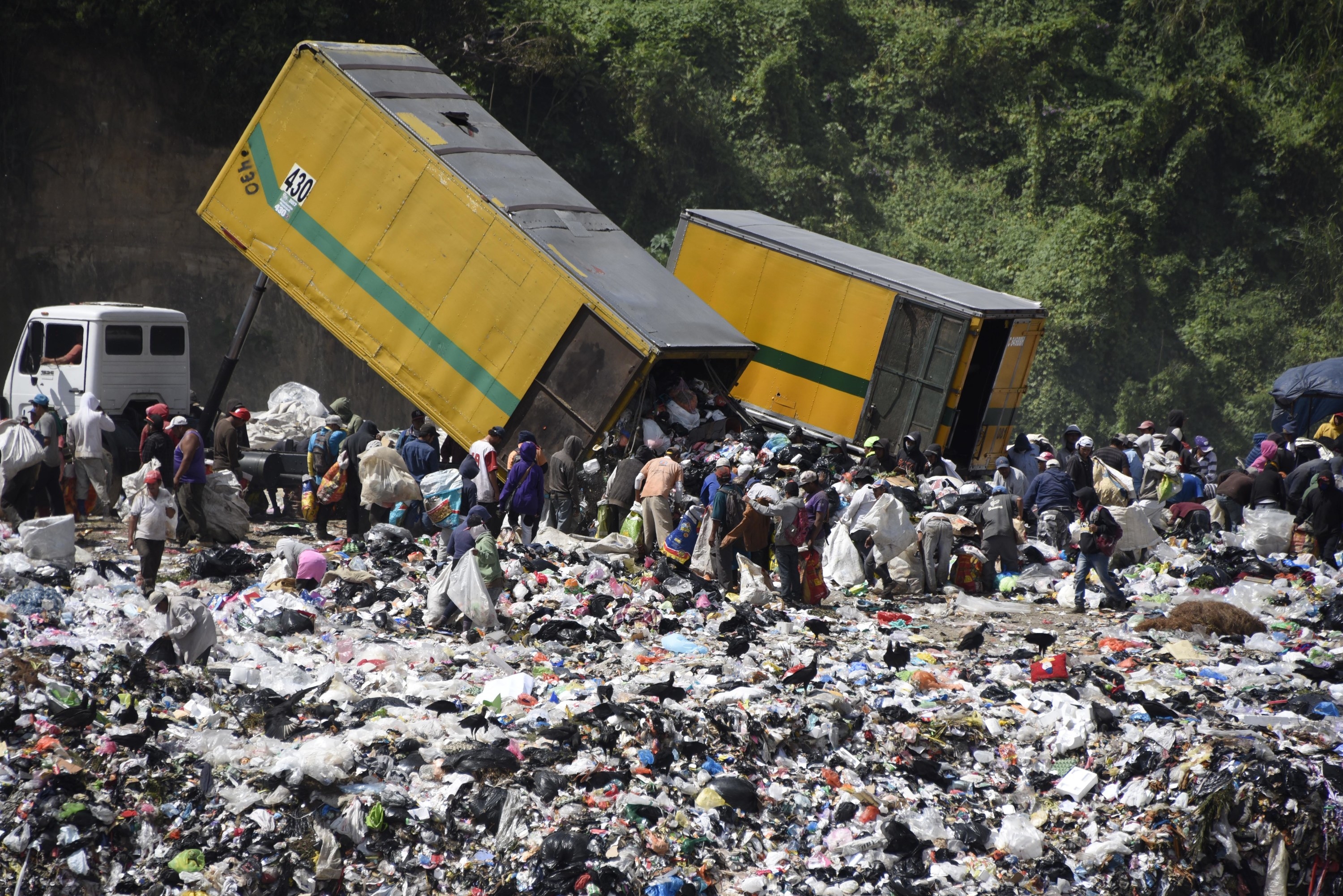 Dozens of people wait as trash trucks empty waste at a landfill in South America