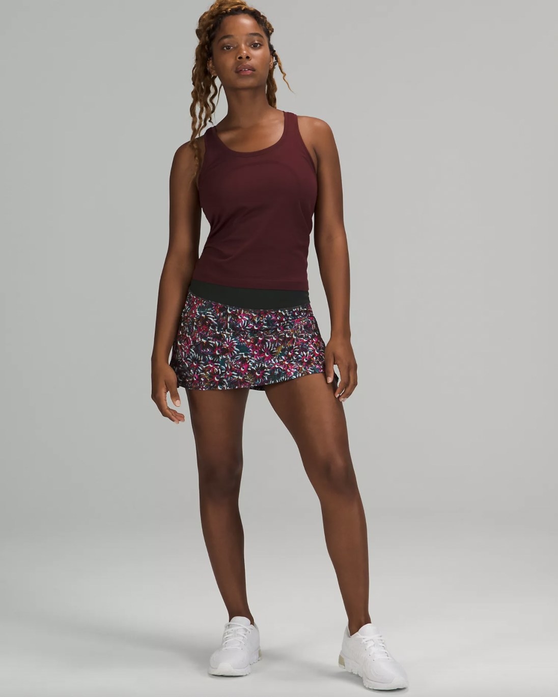A woman wearing a burgundy tank top, a floral skirt, and white sneakers