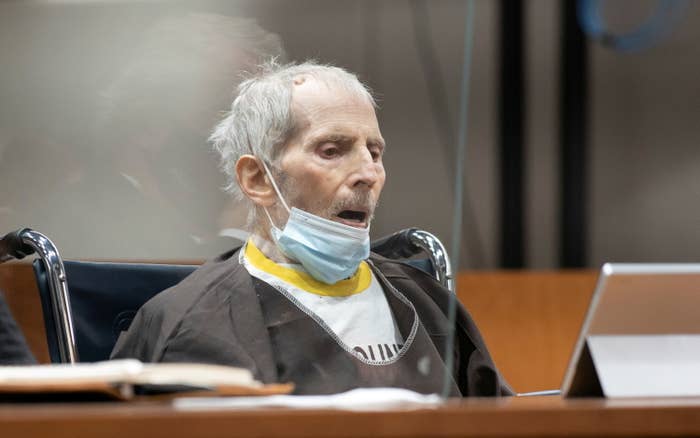 Robert Durst, sitting with his mouth open and a surgical mask below his mouth on his chin, looks down