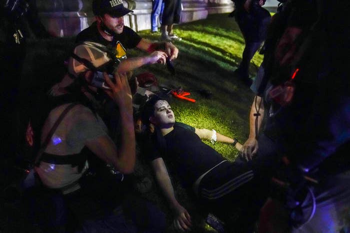 A person lies on the grass with eyes closed as another person in the surrounding crowd tends to them