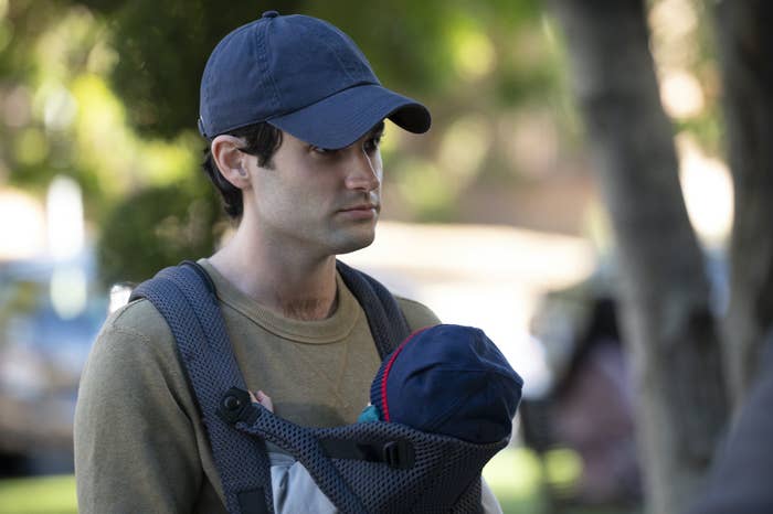 Penn Badgely as Joe; he is wearing a cap and holding Henry in a baby holder