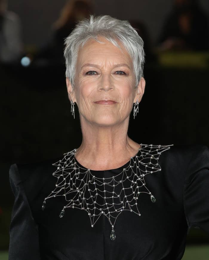 Jamie Lee Curtis smiles for the camera while wearing earrings and a black dress with a white neck design