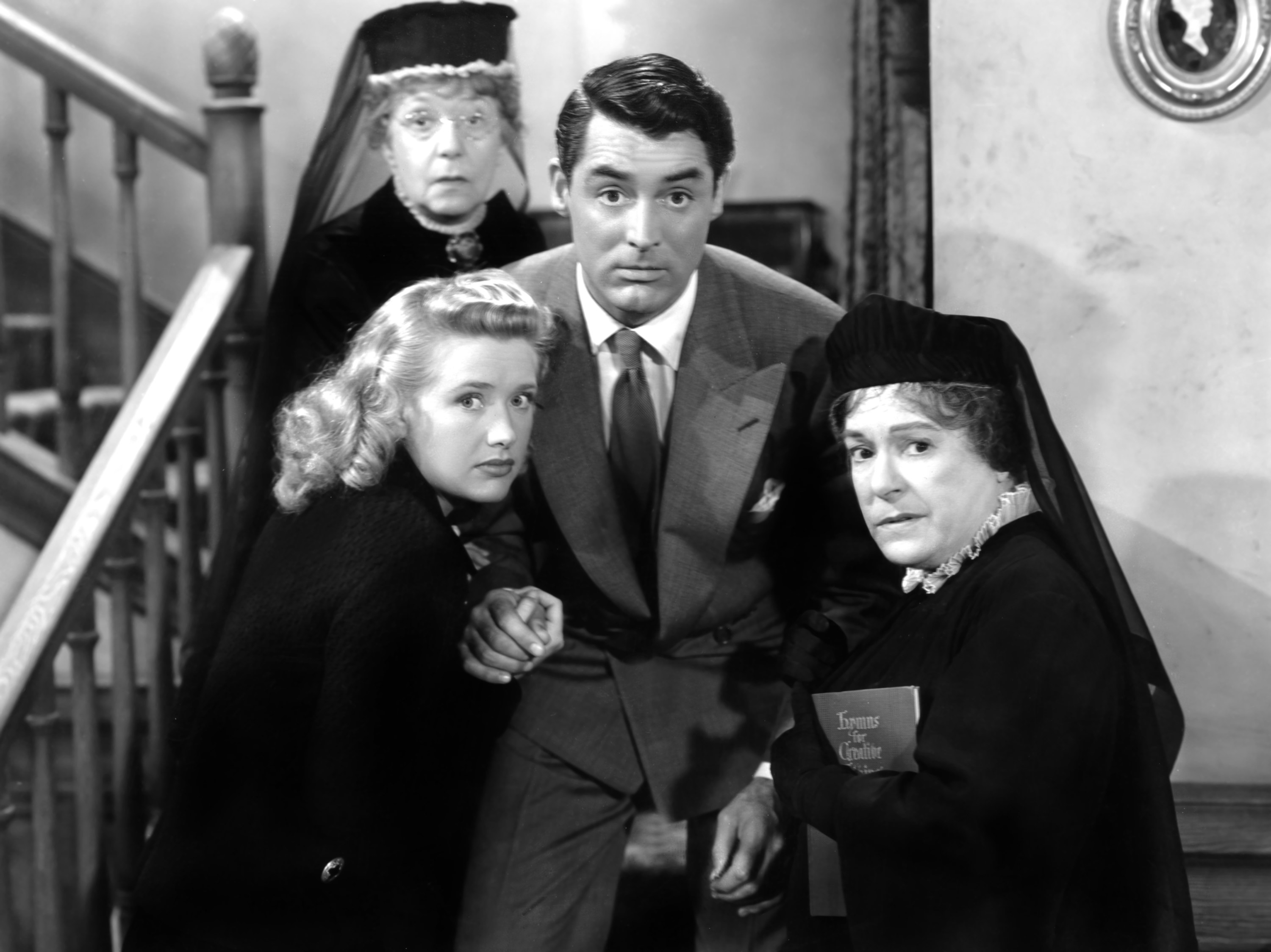 Carey Grant looks into the camera with Priscilla Lane, Jean Adair, and Josephine Hull surrounding him