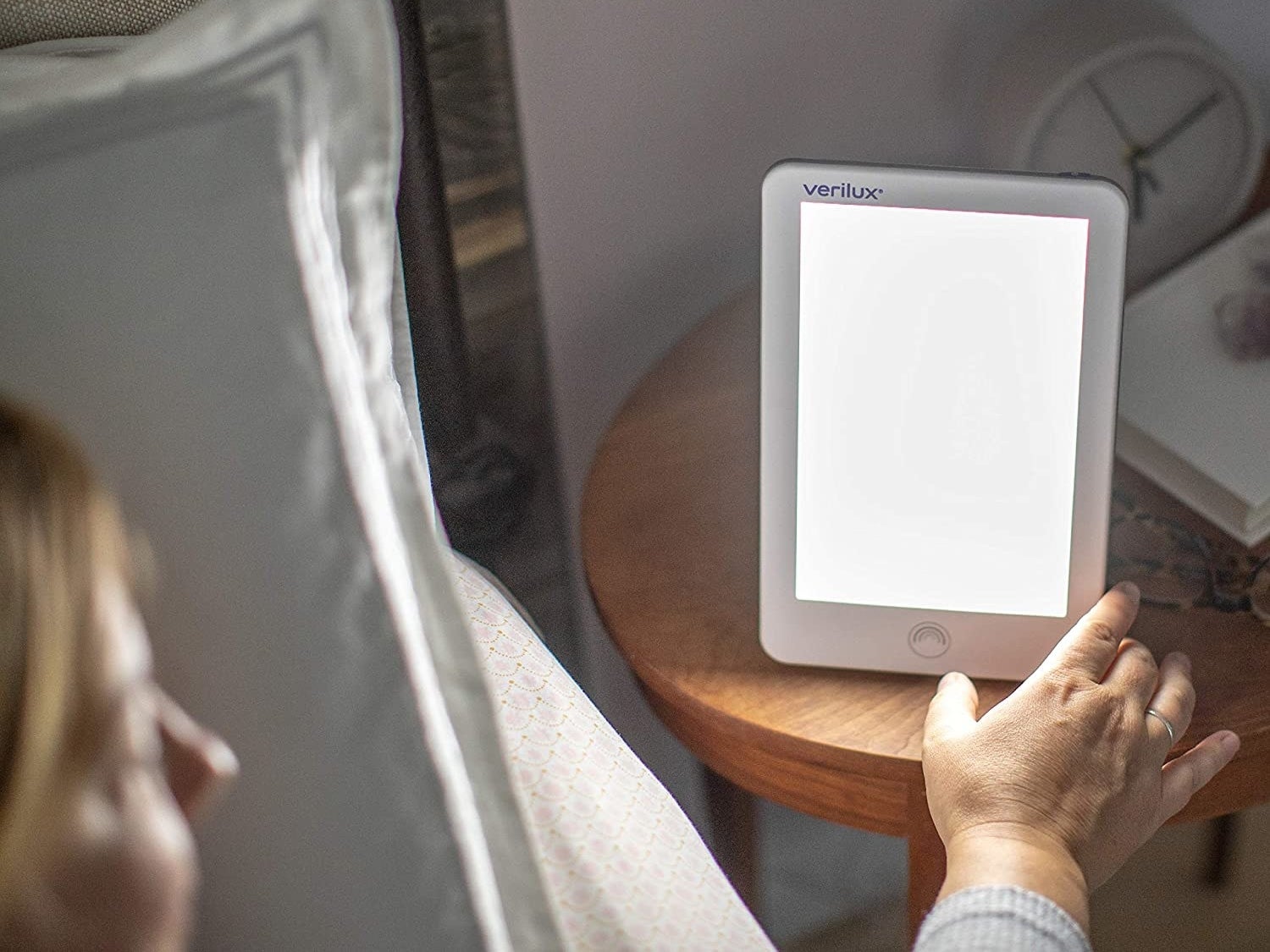 The light positioned upright on a nightstand; someone is reaching out to adjust the brightness