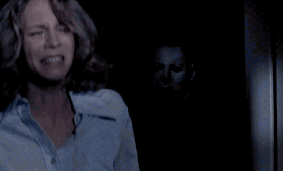 Laurie standing scared in the light as Michael stands in the dark and only half of this mask is visible