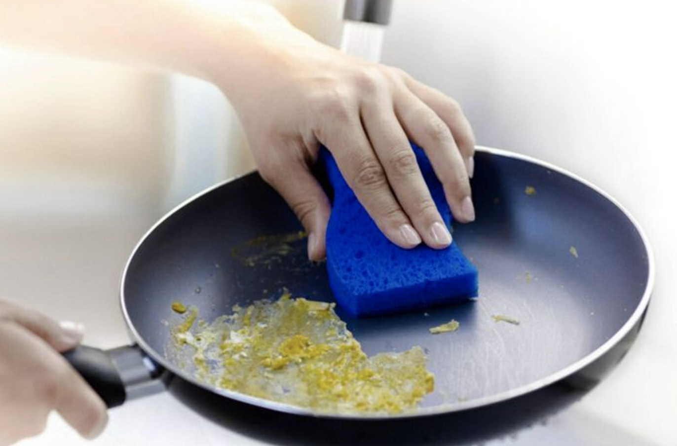 hand uses blue Scotch-Brite sponge to clean egg remains from pan