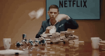 Man with Netflix sign behind him throws out stacks of cash that are on the table in front of him