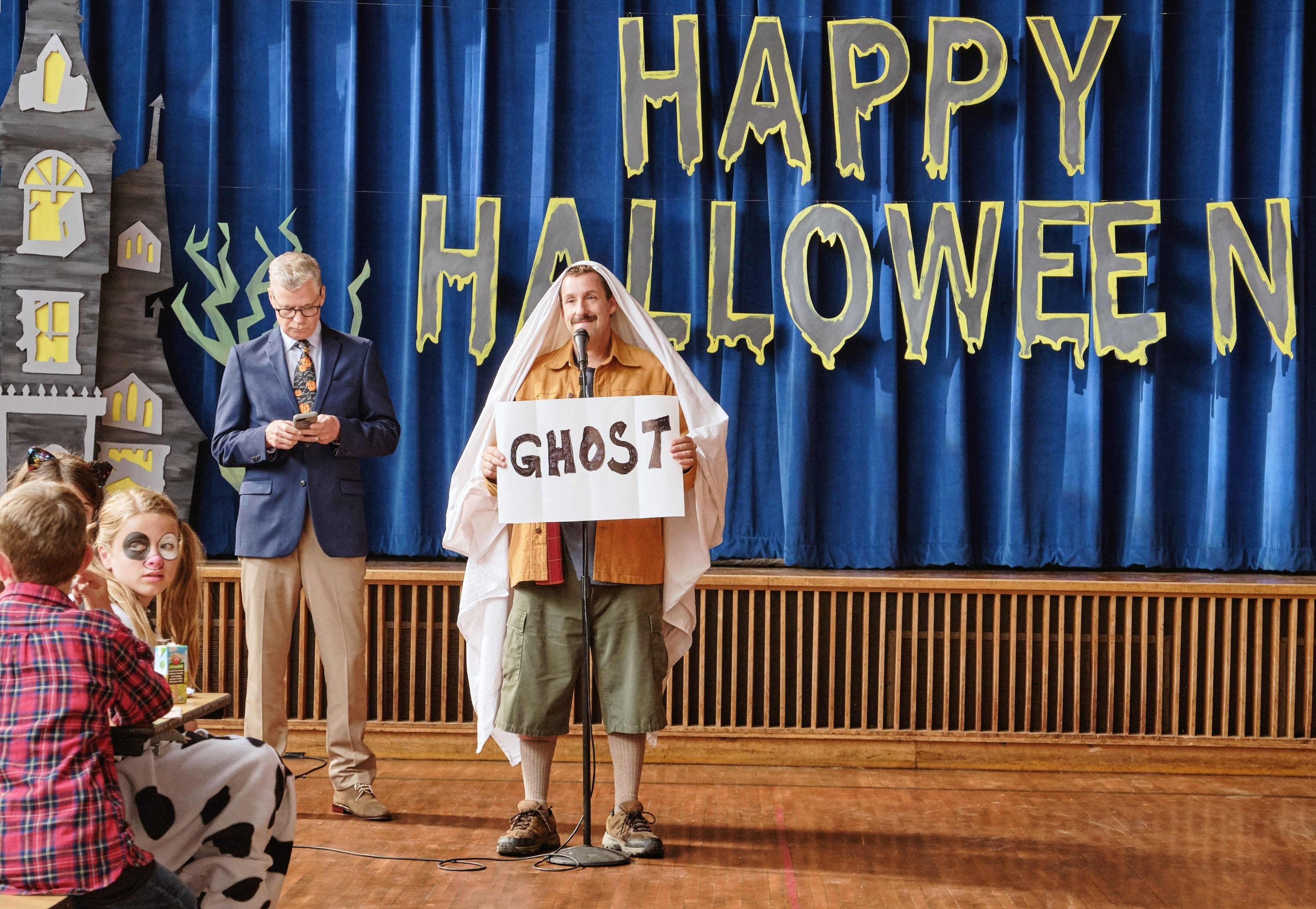 Adam Sandler smiling while holding up a sign that says &#x27;ghost.&#x27;