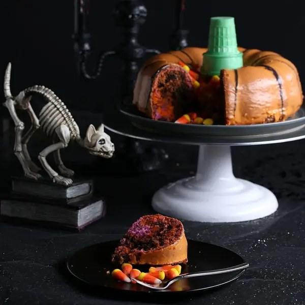 slice of Bundt cake and candy corn on a serving plate, along with a platter of cake to the right and a skeleton cat to the left