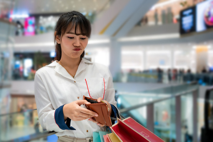 Unhappy person looking in open wallet with disappointed expression while holding color shopping bags at mall. Young model spent too much money during shopping.