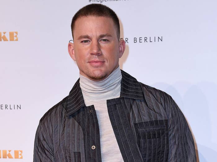 Channing stands on a red carpet in a grey turtleneck and striped button down