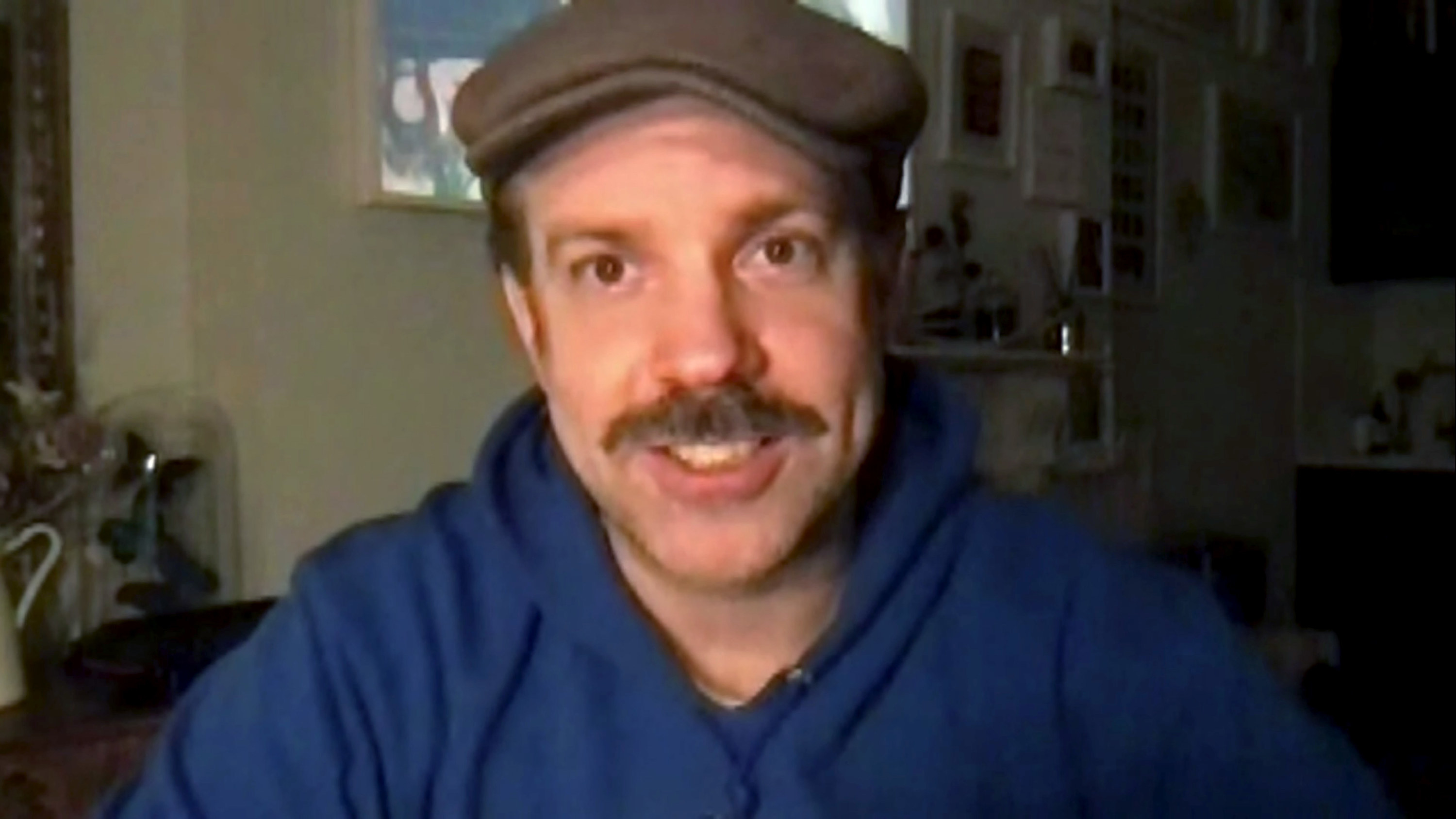Sudeikis looks into a camera on Zoom while wearing a newsboy hat and sweatshirt