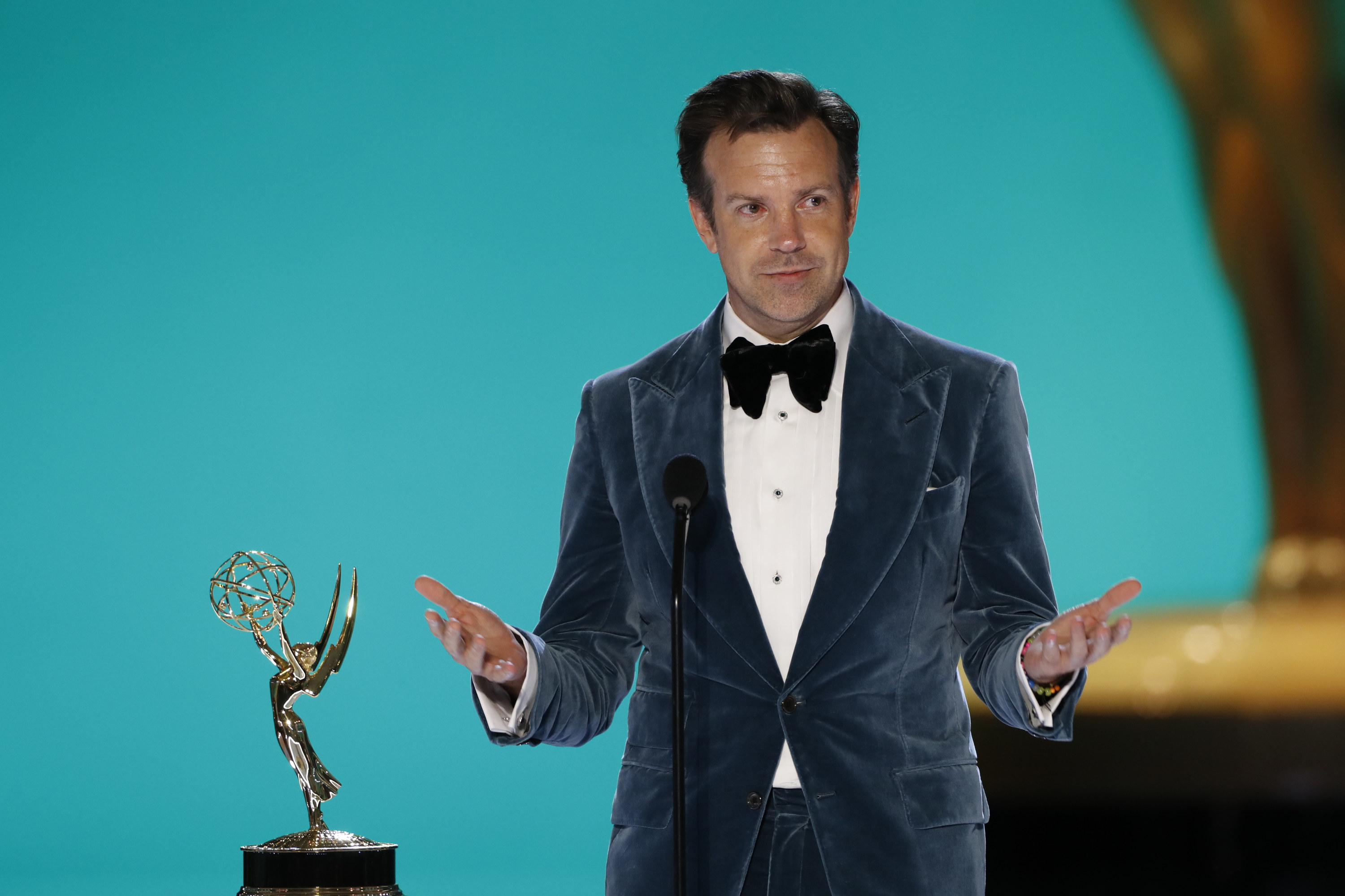 Sudeikis stands on stage next to an Emmy with his arms out