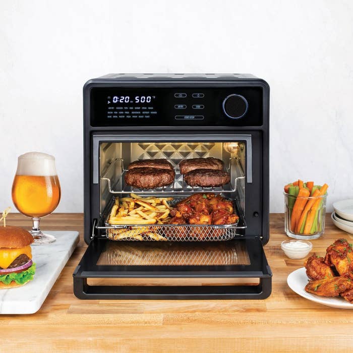 toaster oven with fries, burgers and chicken wings inside. outside on the kitchen counter there's a burger, beer and carrot sticks.