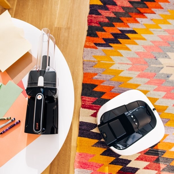 Product image of black and white robot vacuum on carpet and black handheld vacuum on top of a white table