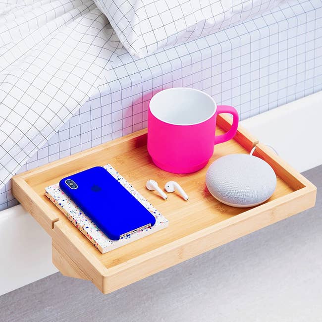 the wood shelf perched on the side of a bed holding a mug, smart home device, earbuds, a phone, and a journal