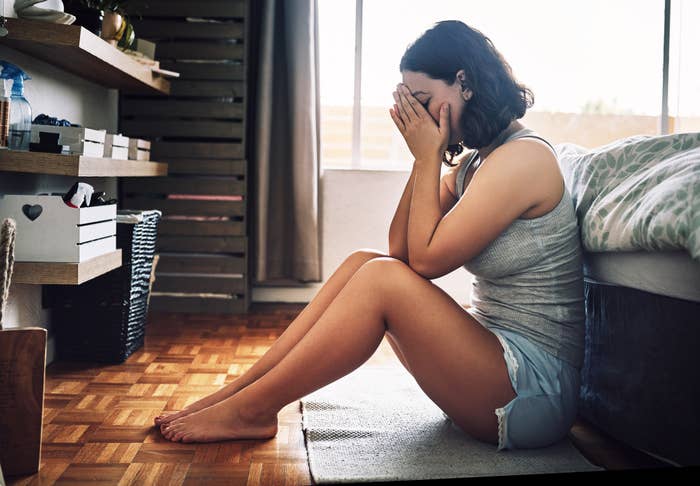 A young woman sitting on the floor beside her bed with her face in her hands