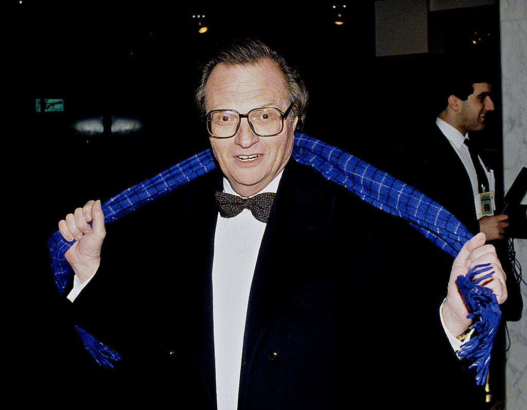 Larry King stretching a scarf behind his neck while smiling for the camera