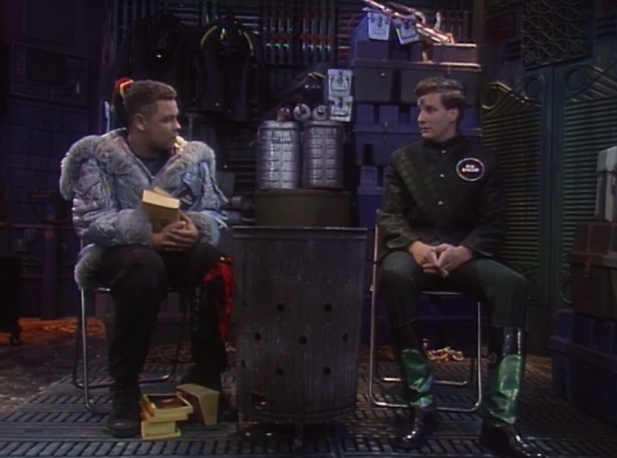 Lister and Rimmer sit in a spaceship in front of a trash can; they&#x27;re looking at each other; Lister holds a book with more books at his feet