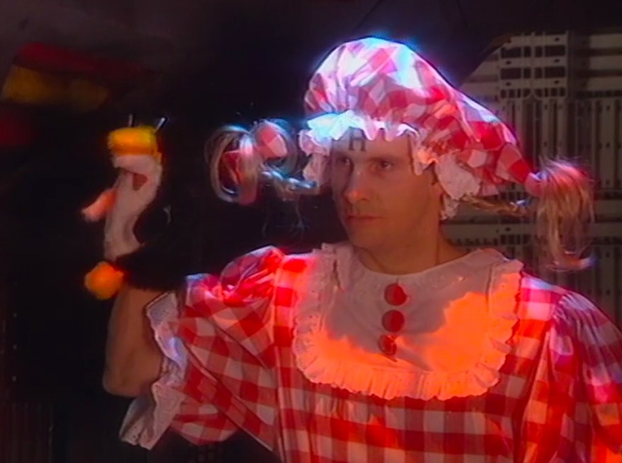 Rimmer wears a gingham dress and bonnet with fake pigtails and has a penguin puppet on his hand