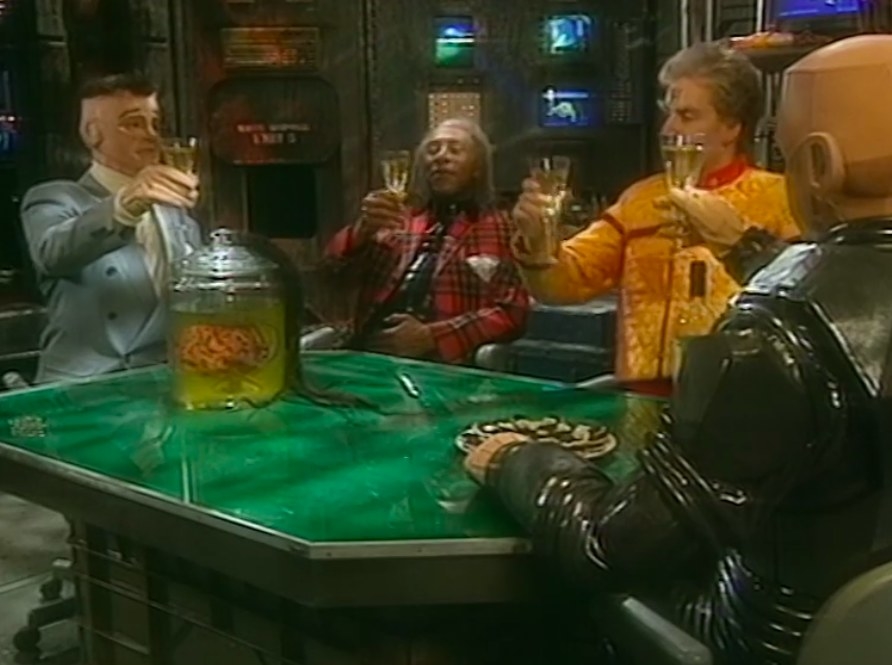 Kryten toasts with alternate reality/older Kryten, The Cat and Rimmer; a brain in a jar sits on the table in front of them