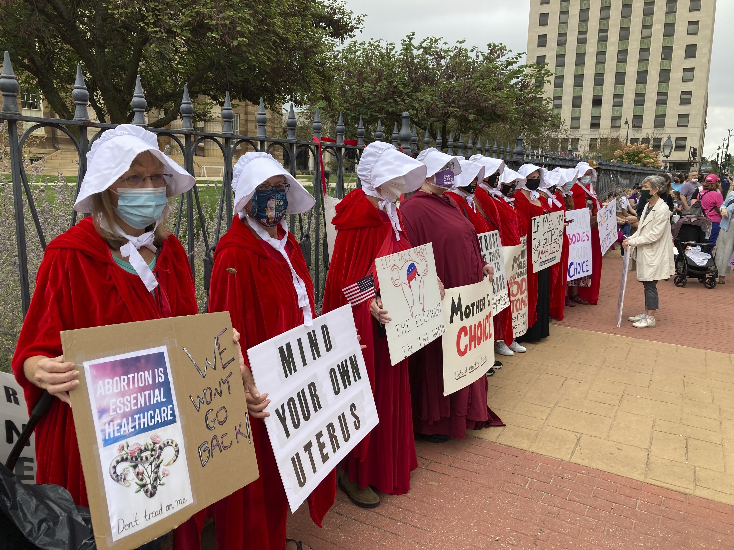 People standing by a gate and dressed as Handmaids from &quot;The Handmaids Tale&quot; hold up signs, including &quot;Mind Your Own Uterus&quot;