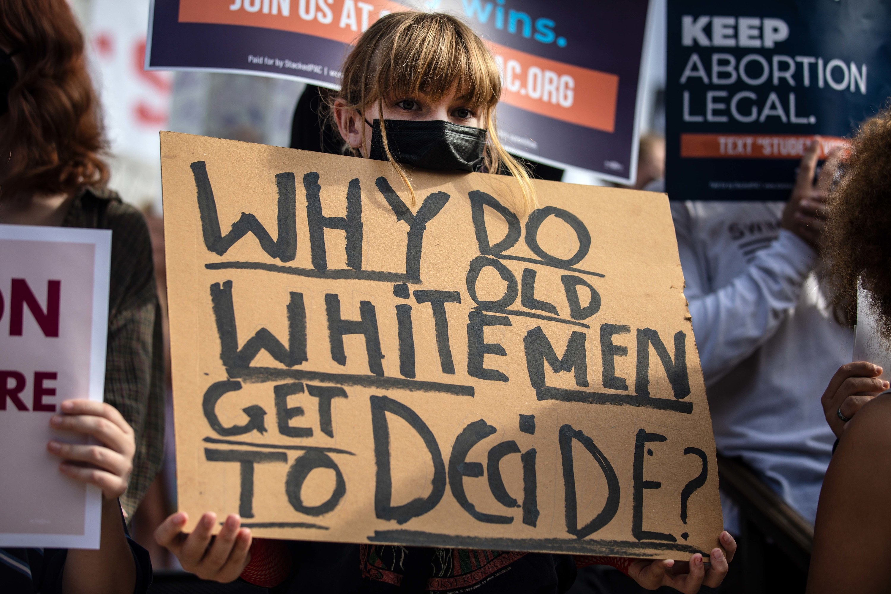 A person holds a sign that reads &quot;Why Do Old White Men Get to Decide?&quot;