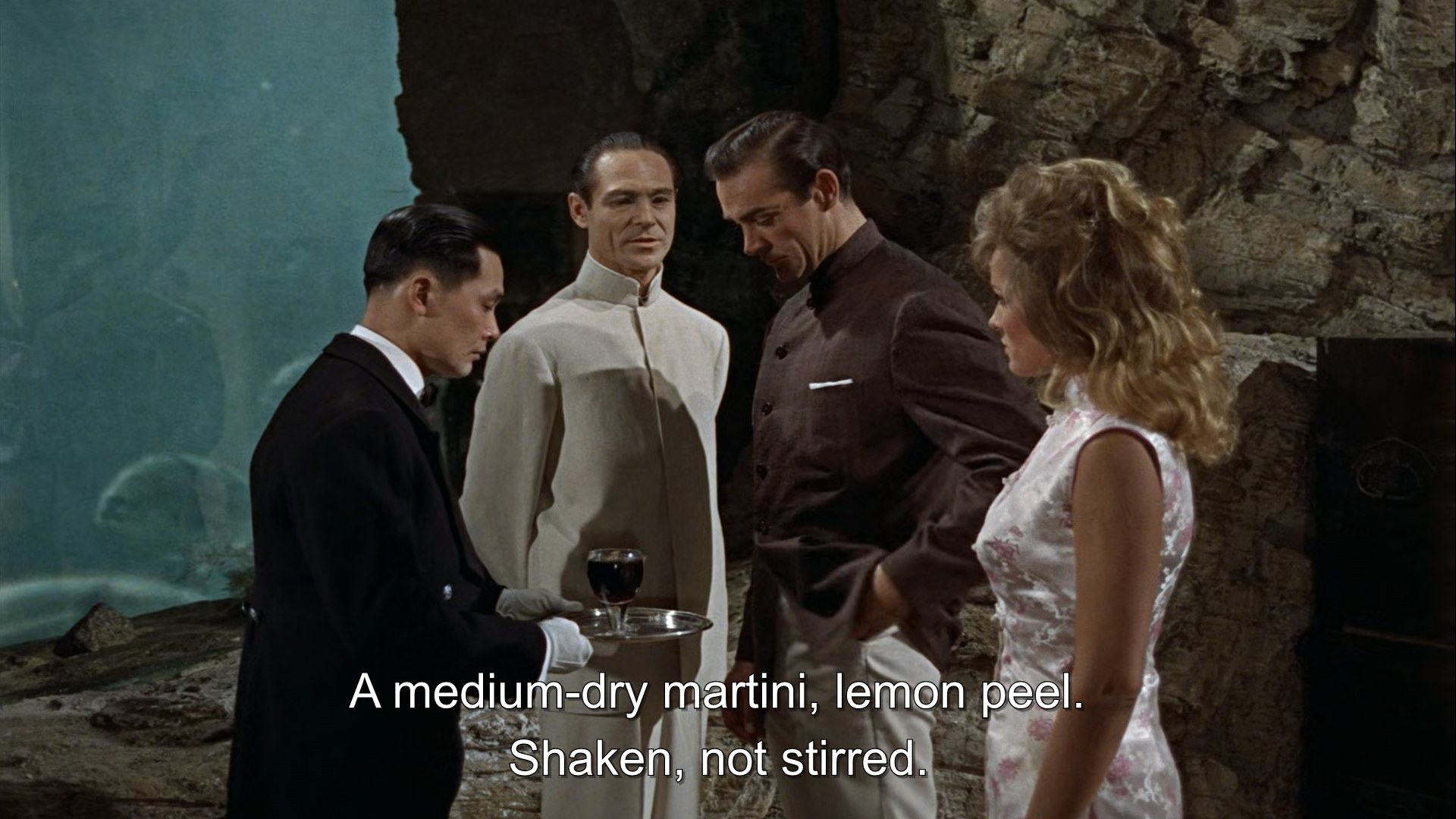 A man serves a cocktail to James Bond while Dr No comments on the contents of the cocktail as Honey Ryder looks at him