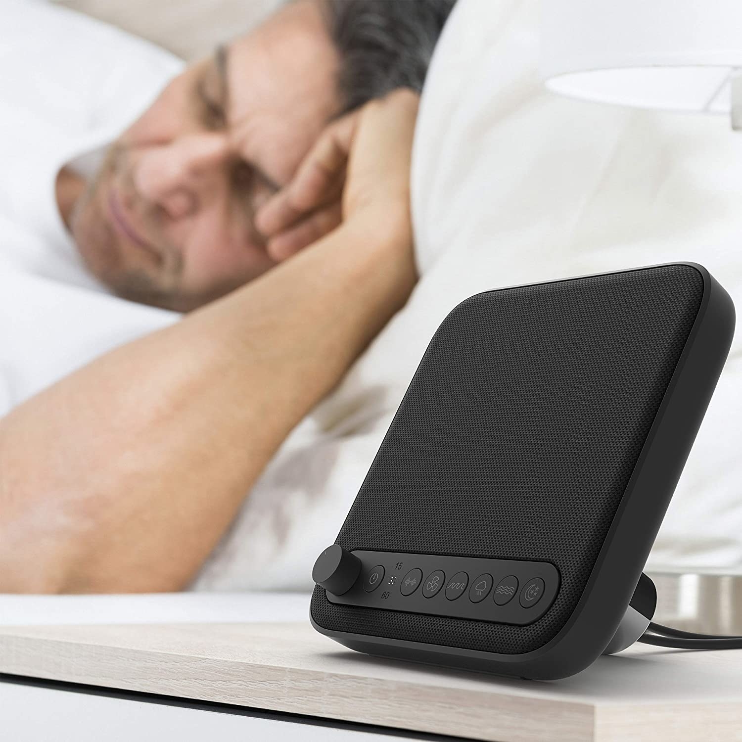 a slim sound machine on a nightstand; a sleeping person is visible in the background