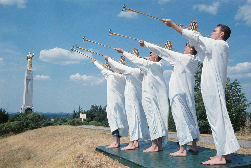 A row of men in matching robes playing wind instrument on a platform with a statue in the background