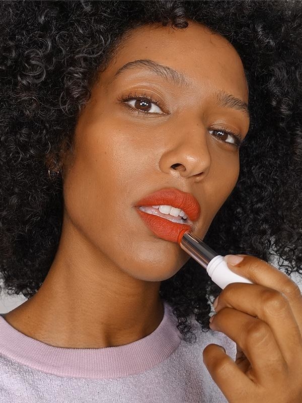 model swipes Glossier Generation G red-orange lipstick on lips