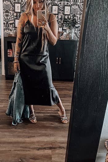 A reviewer posing in the mirror holding a denim jacket while wearing the black silk dress and flip flops.
