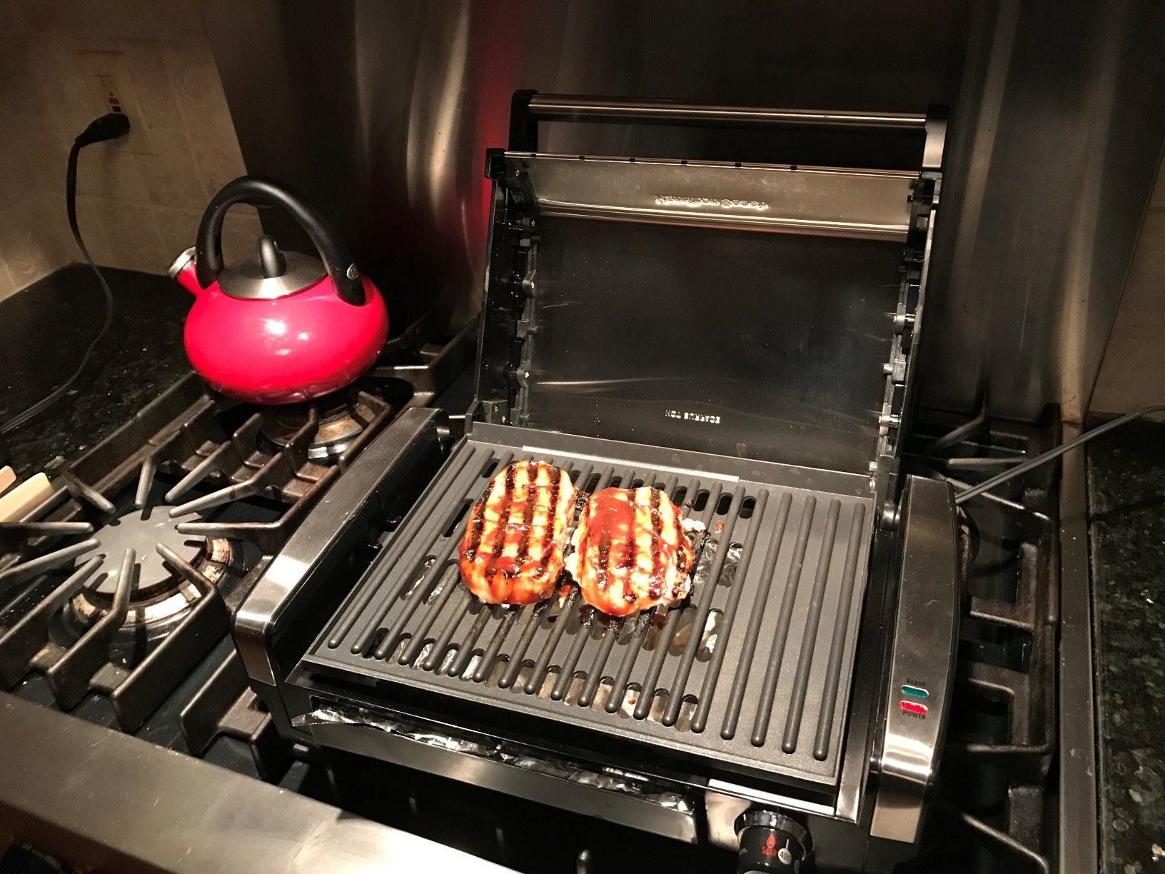 reviewer image of the grill on top of a stove with two piece of meat grilling on top and a red kettle in the background