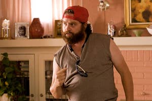 Galifianakis points to himself while wearing a tank shirt, backwards hat, and sunglasses in a living room