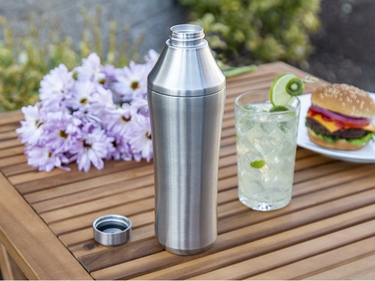 An insulated cocktail shaker on a table with a drink and food.