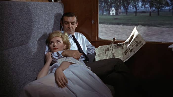 James Bond is reading a newspaper in the compartment of a train while a woman relaxes next to him with his right arm around her