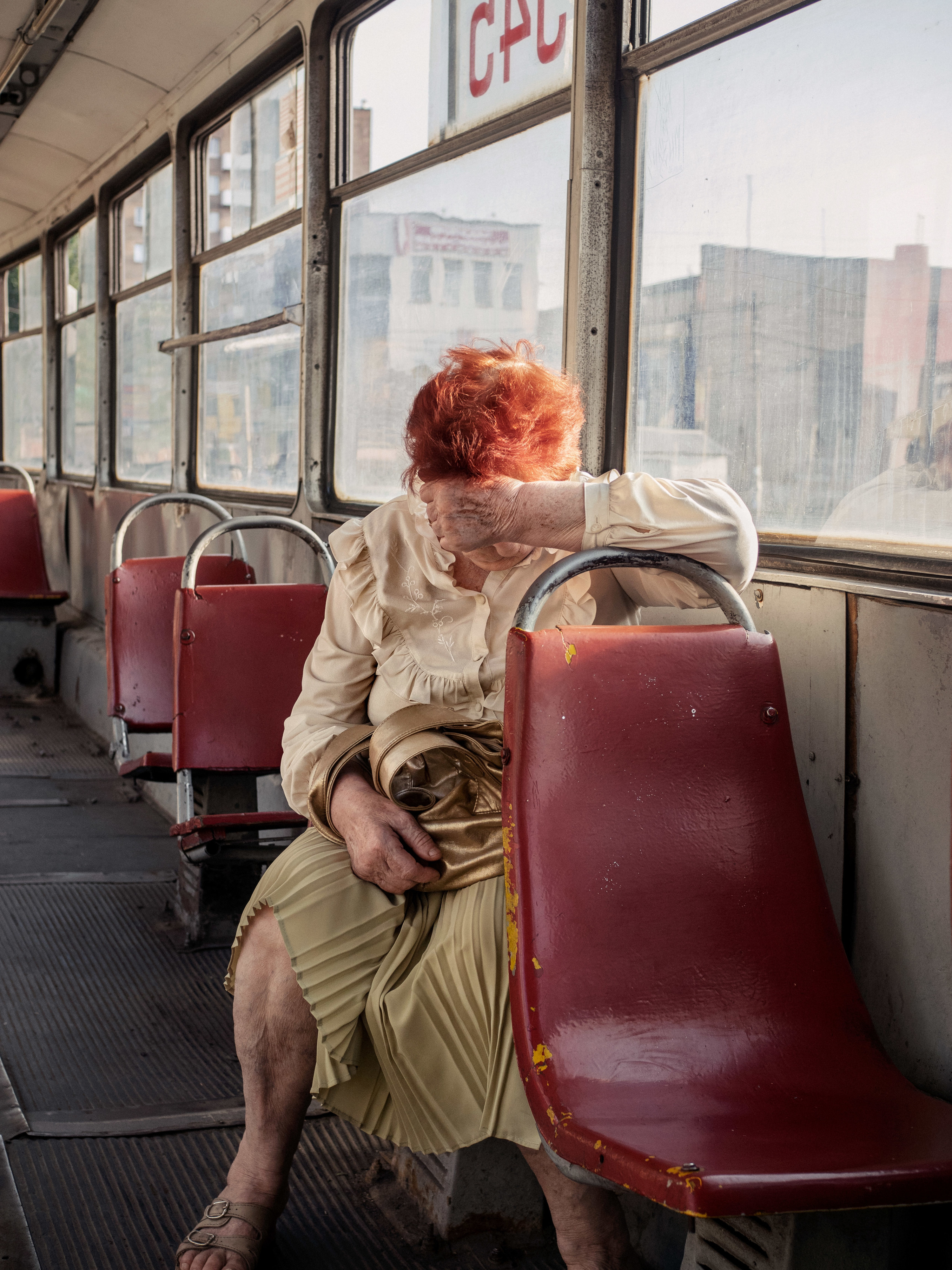 An older person covering their eyes at they sit on a run-down bus