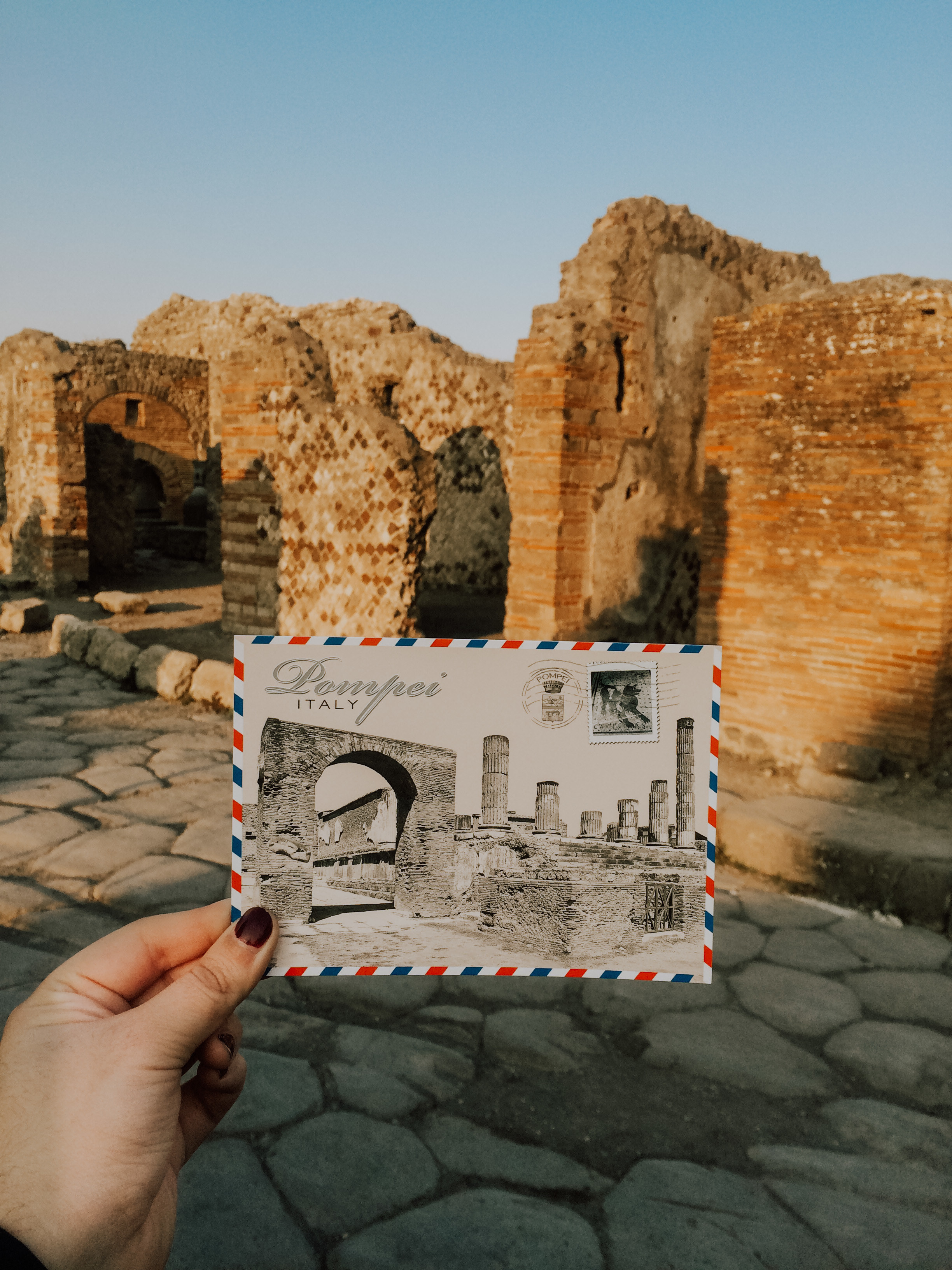 photo of a hand holding up a postcard of Pompeii in front of the ruins in Pompeii