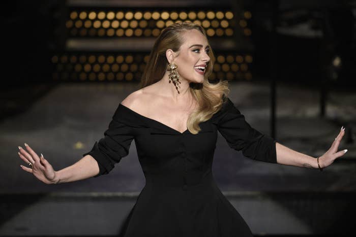 Adele holds her hands out while standing on stage in a black dress
