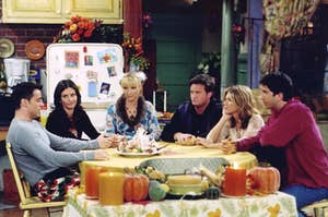 The cast of "Friends" are sitting at a dinner table with Thanksgiving dinner spread