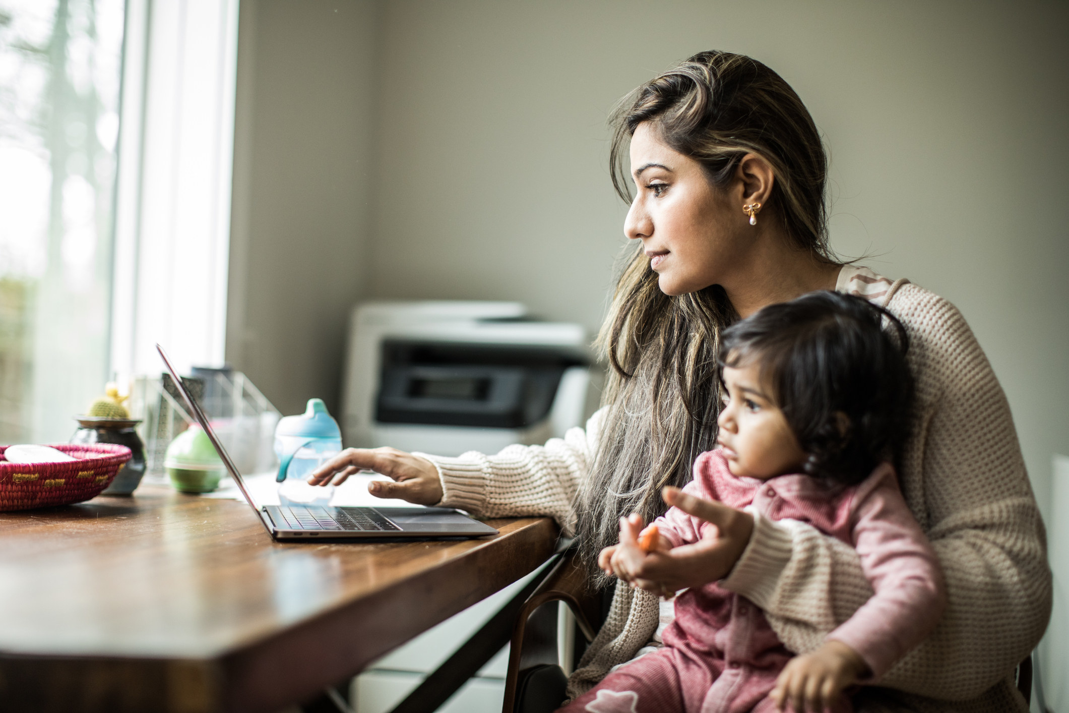 A woman sitting at her desk with her laptop and her baby in her arms
