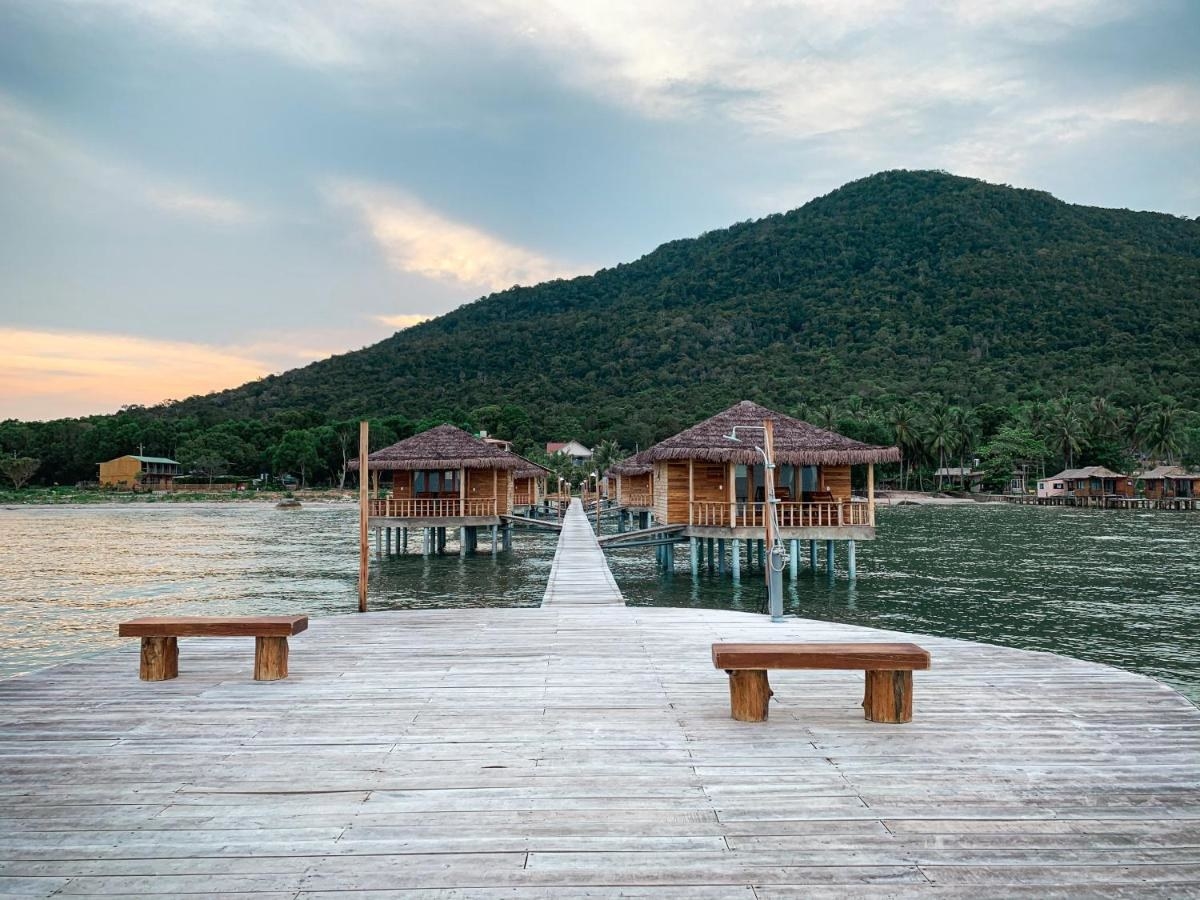 Wooden bungalows over the water in Vietnam with a forested mountain set behind it.