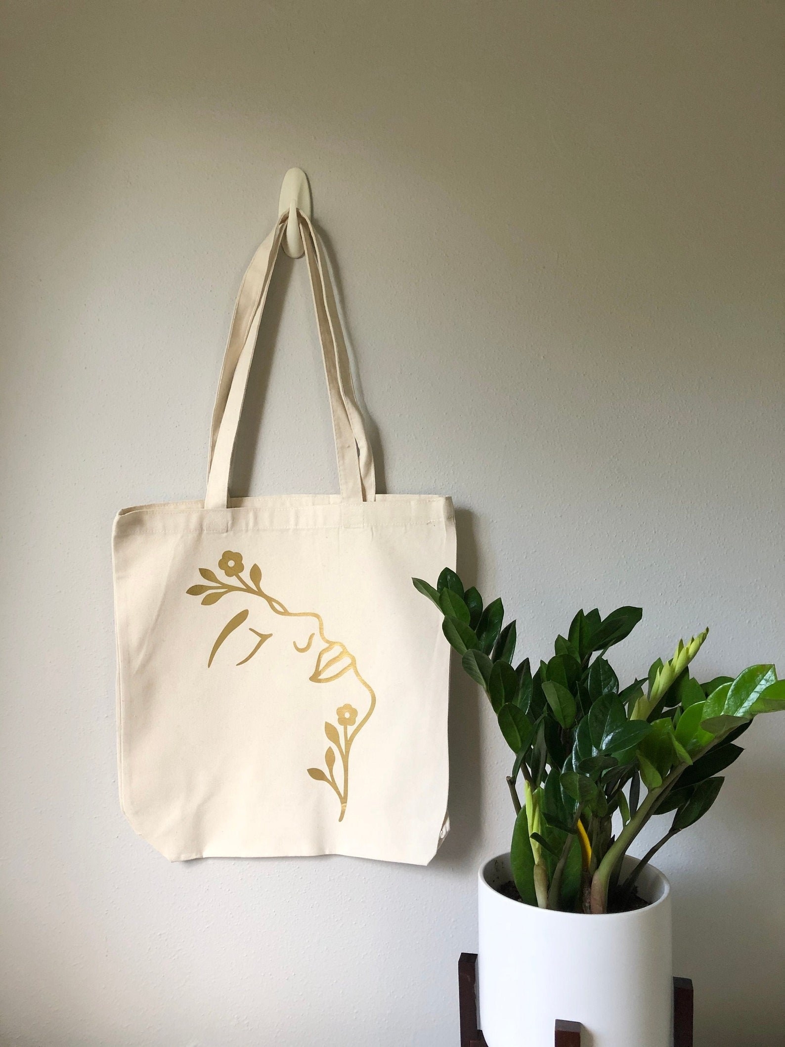 A white tote bag with a gold design featuring a woman&#x27;s face and flowers