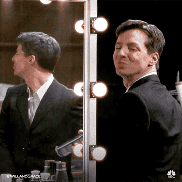 Jack from Will and Grace standing in front of a mirror in a suit spraying loads of hairspray, then turning to the camera and smirking.