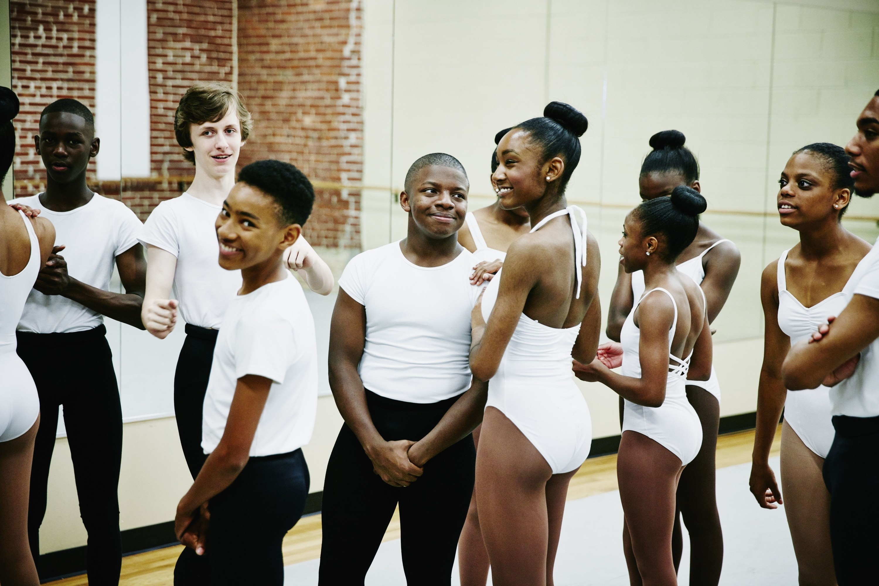 Male and female dancers standing together smiling, girls in white leotards and boys in white tops and black leggings.