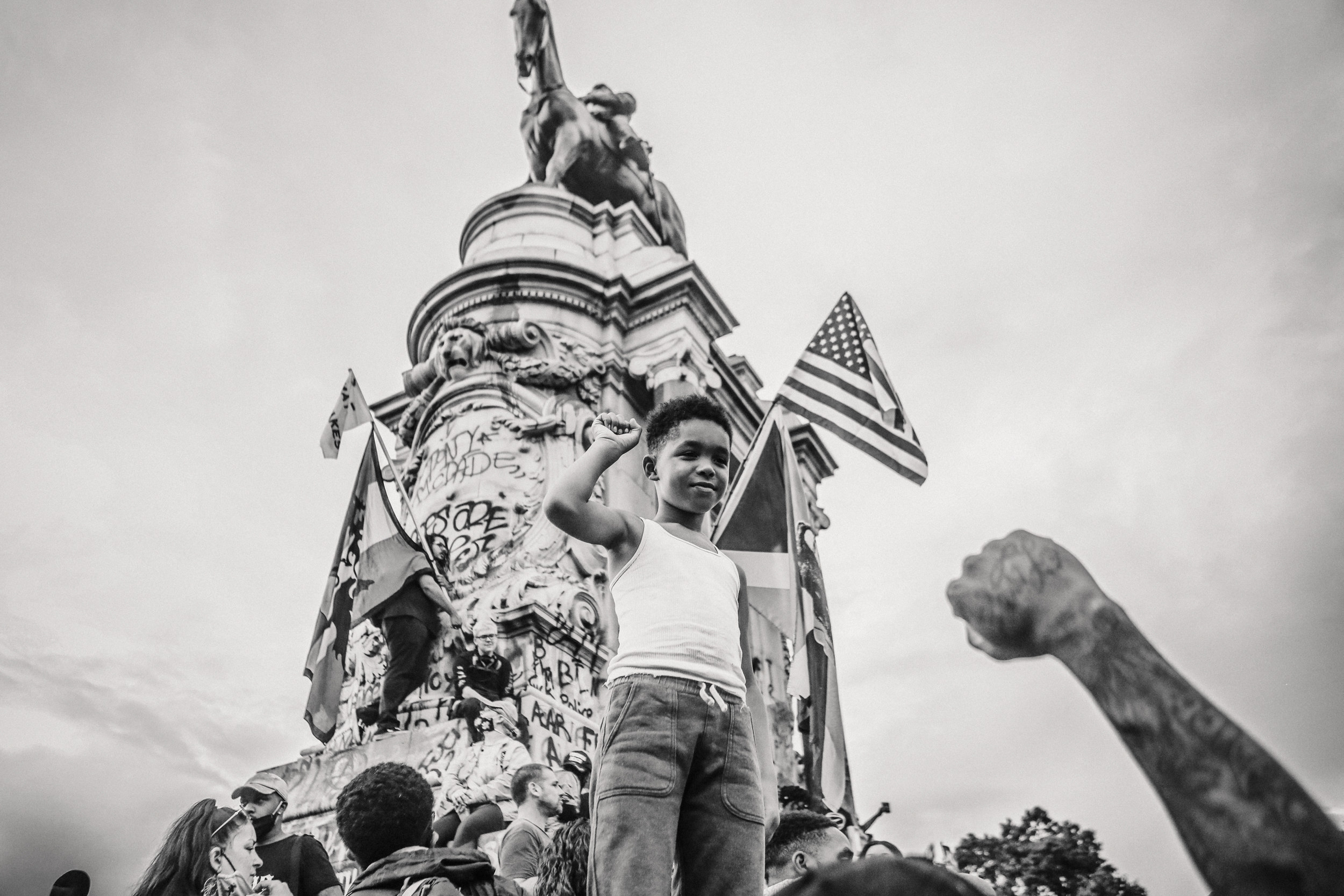 A young boy holds his fist up underneath a statue covered in graffiti 